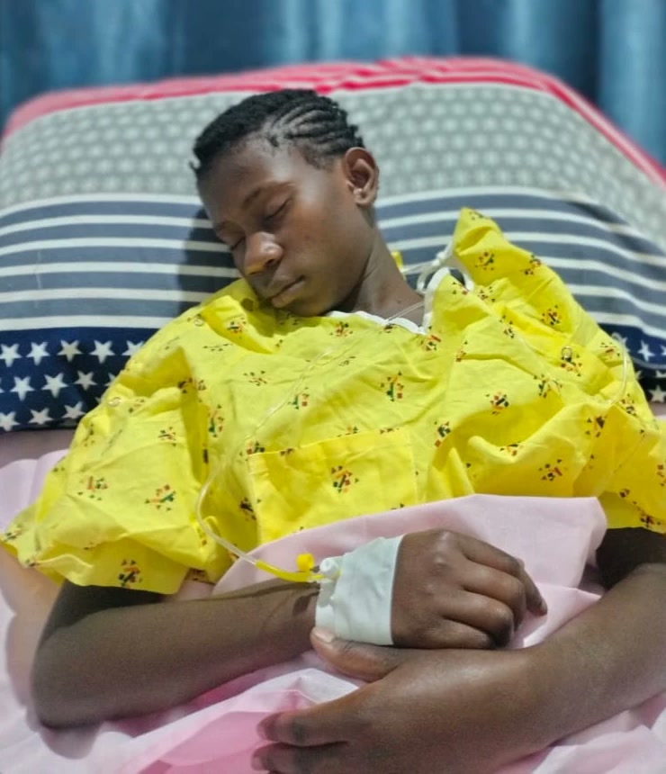 Moreen asleep in a hospital bed after her surgery, in a yellow patterned gown, her hands folded together with a dressing taped over the cannula in her wrist.